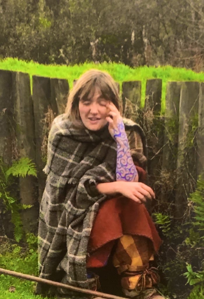 Person with woad painted on arm, sitting next to a wooden palisade wall, wearing a grey woollen cloak, tunic and wool trousers.